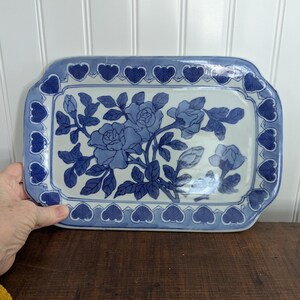 May include: Rectangular ceramic serving tray with a blue and white floral design. The tray features a border of blue hearts and a central motif of blue roses and leaves. The tray is held by a hand and rests on a wooden surface.