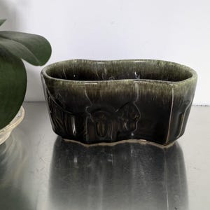 May include: A dark green ceramic planter with a wavy rim and embossed floral designs. The planter is made of glazed pottery and has a rustic, vintage look.