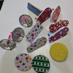 May include: A collection of vintage Christmas tree light reflectors and decorative toppers. The toppers feature various patterns, including stars, dots, and geometric designs in red, green, yellow, and blue. The reflectors are silver with pink and yellow accents.