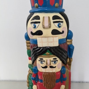May include: A stacked pair of nutcracker figurines. The top nutcracker is wearing a blue and red hat with gold accents. The bottom nutcracker is wearing a red and blue uniform with a green bag.
