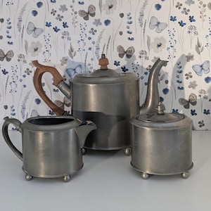 May include: A vintage pewter tea set, including a teapot with a wooden handle, a creamer, and a sugar bowl. The set has a classic design with rounded shapes and small ball feet. The background features a floral wallpaper.