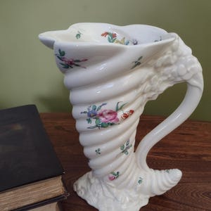 Może przedstawiać: Biały ceramiczny wazon w kształcie muszli z kwiatowym wzorem. Wazon ma uchwyt i szeroki otwór na górze.