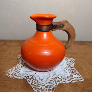 May include: Orange ceramic pitcher with a wooden handle and a copper band around the neck. The pitcher is sitting on a white crocheted doily.
