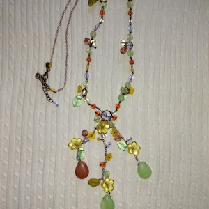 Puede incluir: Un delicado collar con una cadena de color cobre y intrincados diseños florales y de hojas. El collar incorpora detalles de flores amarillas, cuentas verdes y naranjas, y colgantes en forma de lágrima en tonos verdes y marrones.