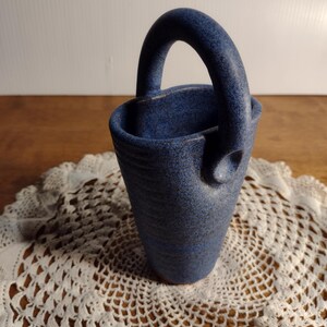 May include: A small blue ceramic vase with a handle. The vase has a speckled blue glaze and a rounded bottom.