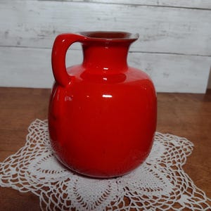 May include: A bright red ceramic pitcher with a brown rim and a handle. The pitcher is sitting on a white crocheted doily.