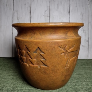 Könnte beinhalten: Ein brauner Keramik-Pflanztopf mit breitem Rand und leicht konischem Körper. Der Topf hat geschnitzte Designs von Kiefern und einem Elch. Der Hintergrund ist eine helle Holztafel.