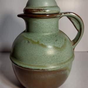 May include: A green and brown ceramic pitcher with a lid. The pitcher has a round body and a handle. The lid has a knob on top.