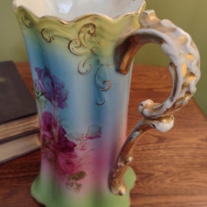 Puede incluir: Una jarra de porcelana vintage con un esmalte degradado verde, azul y rosa. La jarra presenta un diseño floral con detalles dorados y una asa decorativa.