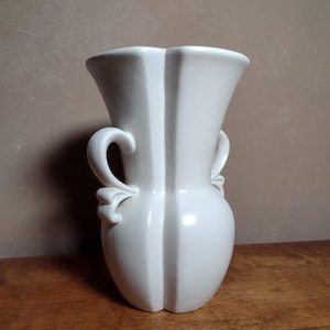 Puede incluir: Un jarrón de cerámica blanco con dos asas. El jarrón tiene un acabado liso y brillante y un diseño simple y elegante.