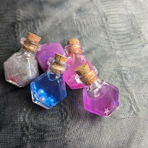 Puede incluir: Cuatro pequeñas botellas de vidrio con tapones de corcho llenas de purpurina colorida y cuentas iridiscentes. Las botellas están dispuestas en un grupo sobre una superficie gris texturizada.