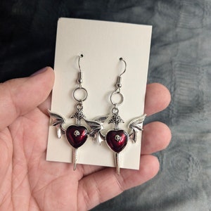 May include: A pair of silver dangle earrings with red heart-shaped charms. Each charm has a skull design and is topped with a pair of wings.