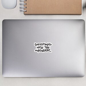 May include: A silver laptop computer with a black and white sticker that says "QUESTIONS ARE THE ANSWER" on the lid. The laptop is on a white surface with a white mouse, a brown notebook, and two small potted plants.