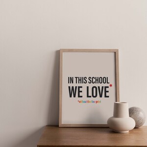 以下が含まれることがあります： 「In this school we love* *without the fine print」というテキストが書かれた額装されたプリント。テキストは黒で書かれており、赤いアスタリスクが付いています。「without the fine print」というテキストは虹色です。プリントは薄いベージュの背景に印刷され、薄い木製のフレームで額装されています。プリントは木製のテーブルの上に置かれています。