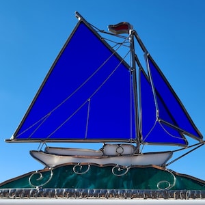 May include: A stained glass sailboat with a blue sail and a white hull. The sailboat is sitting on a green base with a wave design.