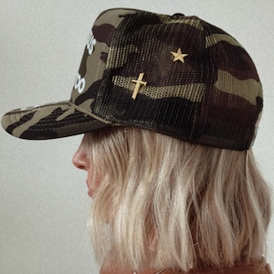 Puede incluir: Gorra de béisbol de camuflaje con parte trasera de malla, alfileres dorados de cruz y estrella. La gorra tiene el texto "BUSY AS FUCK" en la parte delantera. La persona lleva una camisa marrón.