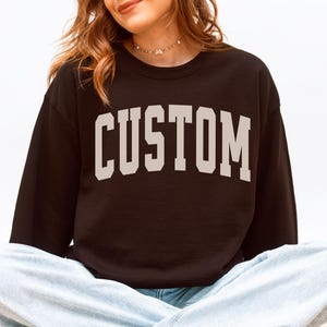 May include: A black sweatshirt with the word "CUSTOM" printed in a light beige, collegiate-style font. The sweatshirt is long-sleeved and has a crew neck. The person is wearing light blue jeans and white sneakers.