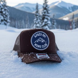 May include: A brown trucker hat with a mesh back and a flat brim, resting in the snow. The hat features a circular patch with the text "Après-Ski Moms Club" and a mountain graphic in navy blue and white.