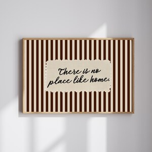 May include: A framed print with a brown and cream striped pattern. The artwork displays the phrase "There is no place like home" in black script on a textured, cream-coloured rectangle.