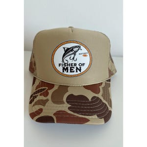 Puede incluir: Gorra de camionero color canela con un patr&oacute;n de camuflaje marr&oacute;n en la visera. La gorra presenta un parche circular con un dise&ntilde;o de pez y anzuelo, y el texto "FISHER OF MEN".