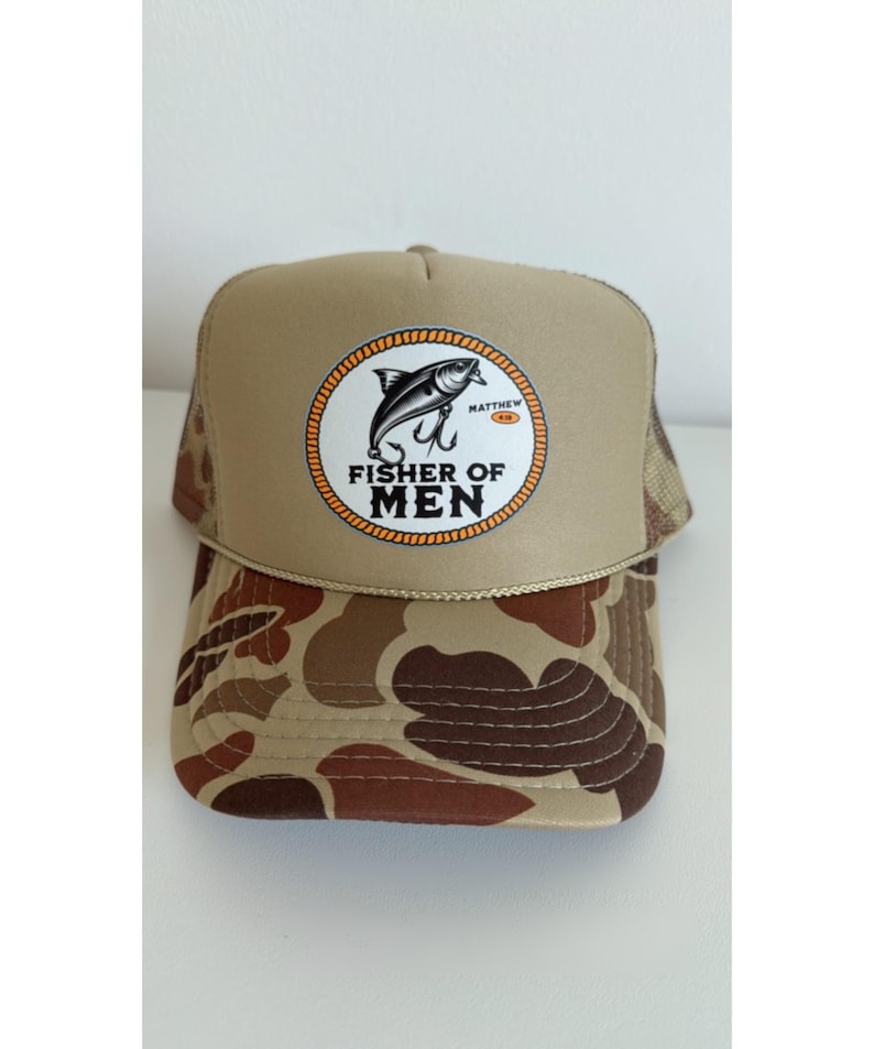Puede incluir: Gorra de camionero color canela con un patr&oacute;n de camuflaje marr&oacute;n en la visera. La gorra presenta un parche circular con un dise&ntilde;o de pez y anzuelo, y el texto "FISHER OF MEN".