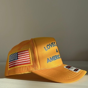 May include: Mustard yellow trucker cap with the text "LOVES & AMERICA" in blue. The cap has a mesh back and a patch of the American flag on the side.