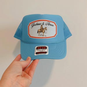 Puede incluir: Gorra de camionero azul claro con parte trasera de malla y visera curva. El panel frontal presenta un parche rectangular con el texto "Yeehaw & Amen" y una ilustraci&oacute;n de un vaquero a caballo. Cierre a presi&oacute;n.