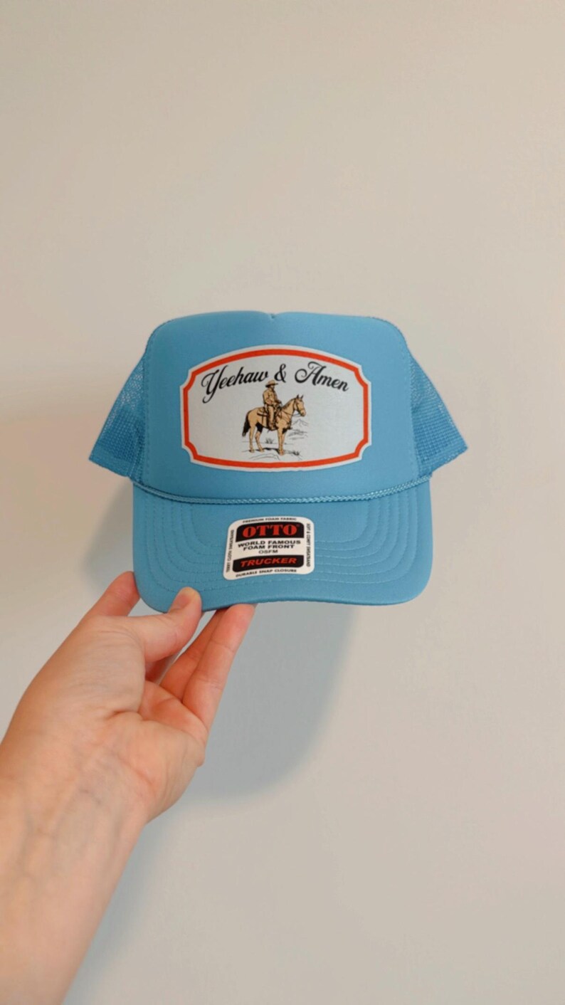 Puede incluir: Gorra de camionero azul claro con parte trasera de malla y visera curva. El panel frontal presenta un parche rectangular con el texto "Yeehaw & Amen" y una ilustraci&oacute;n de un vaquero a caballo. Cierre a presi&oacute;n.