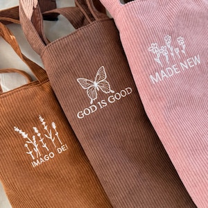 May include: Three tote bags made of brown corduroy with white embroidered designs. The bags feature the phrases "Imago Dei", "God is Good", and "Made New".