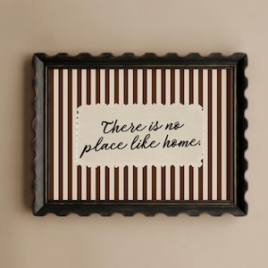 May include: Framed wall art with a decorative border. The artwork has a background of brown and cream vertical stripes. A cream rectangle in the centre displays the text "There is no place like home."