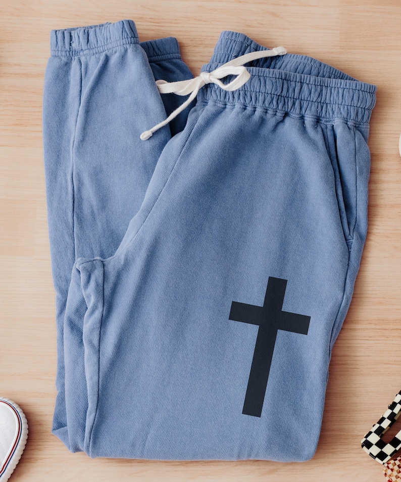 May include: Blue sweatpants with a black cross graphic. The sweatpants have a drawstring waist and a relaxed fit.