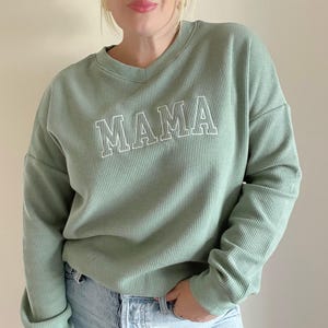 May include: A sage green waffle knit long-sleeve shirt with a white embroidered "MAMA" graphic.