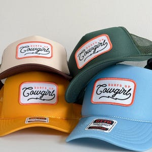 May include: Four trucker hats in different colours: beige, green, yellow, and light blue. Each hat has a rectangular patch with the words "Surfs Up Cowgirl" in a script font. The hats have mesh backs and curved peaks.