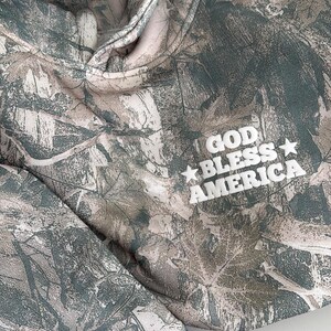 May include: A camouflage-patterned hoodie with the words "GOD BLESS AMERICA" in white block letters. The hoodie features a drawstring hood and a soft, muted color palette of greens, browns, and creams.