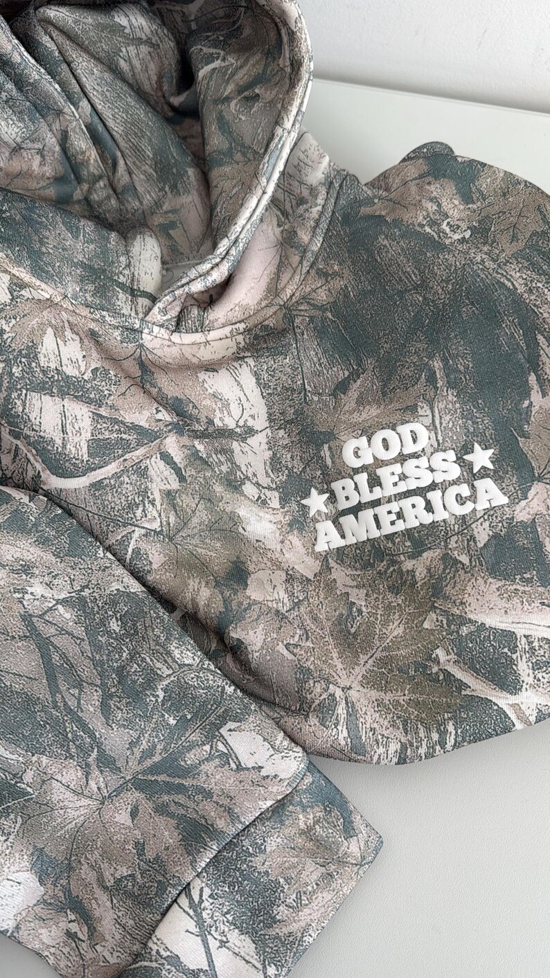 May include: A camouflage-patterned hoodie with the words "GOD BLESS AMERICA" in white block letters. The hoodie features a drawstring hood and a soft, muted color palette of greens, browns, and creams.