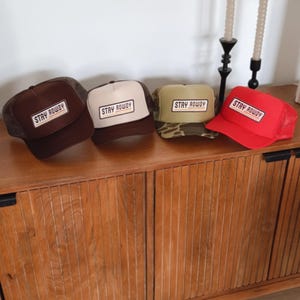 May include: Four trucker hats in brown, tan, camouflage, and red. Each hat features a rectangular patch with the words "STAY ROWDY" in a bold, sans-serif font. The hats are displayed on a wooden cabinet.
