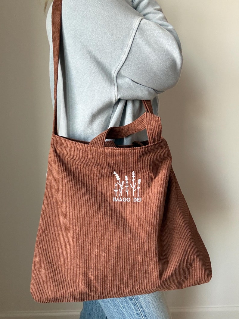 Puede incluir: Una bolsa de mano de pana marr&oacute;n con un dise&ntilde;o bordado en blanco de una planta y el texto "IMAGO DEI".