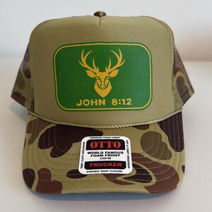 Puede incluir: Gorra de camionero con estampado de camuflaje y parte trasera de malla verde. La parte delantera presenta un rect&aacute;ngulo verde con una cabeza de ciervo dorada y el texto "JOHN 8:12". Cierre a presi&oacute;n.