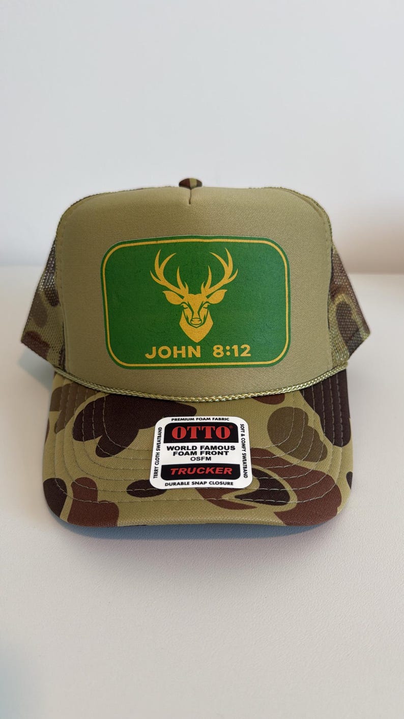 Puede incluir: Gorra de camionero con estampado de camuflaje y parte trasera de malla verde. La parte delantera presenta un rect&aacute;ngulo verde con una cabeza de ciervo dorada y el texto "JOHN 8:12". Cierre a presi&oacute;n.