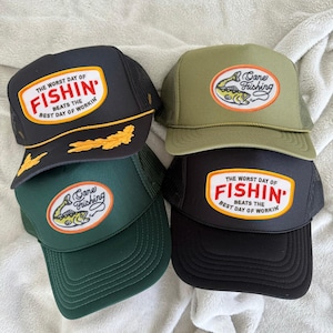 May include: Four trucker hats in black, green, and olive green. Each hat has a mesh back and a front patch with fishing-related text and graphics. The text on the patches includes "The Worst Day of Fishin' Beats the Best Day of Workin'" and "Cane Fishing."