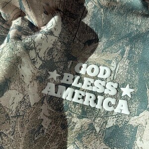 May include: Camouflage-patterned hoodie with the text "GOD BLESS AMERICA" in white block letters and stars. The hoodie features a blend of green, brown, and tan colors, creating a woodland camouflage design.