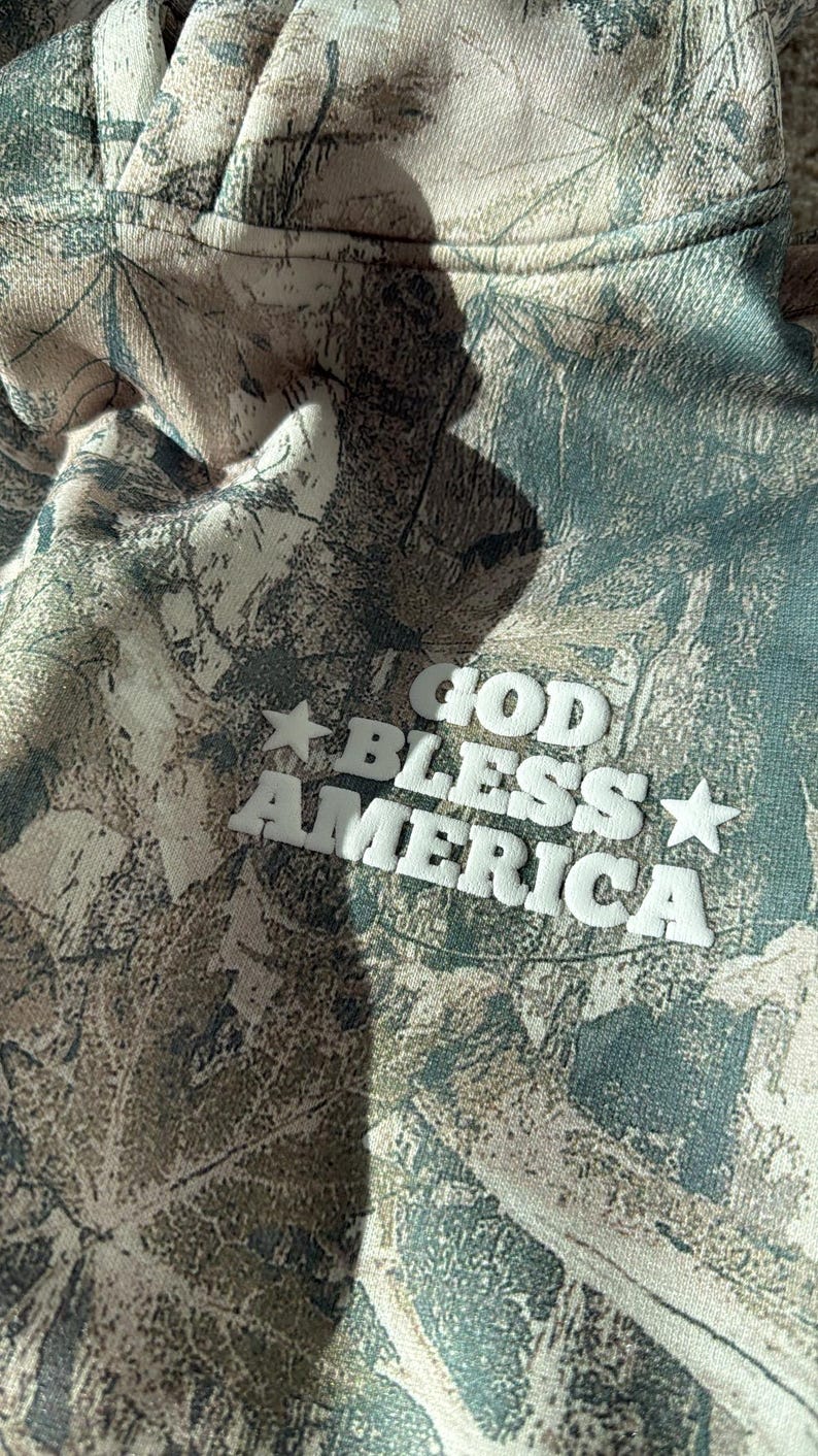 May include: Camouflage-patterned hoodie with the text "GOD BLESS AMERICA" in white block letters and stars. The hoodie features a blend of green, brown, and tan colors, creating a woodland camouflage design.