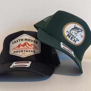 Puede incluir: Dos gorras de camionero. Una gorra negra con un parche con el texto "Faith Moves Mountains" y un gr&aacute;fico de monta&ntilde;a. La otra es verde oscuro con un parche que dice "Fisher of Men" y un gr&aacute;fico de pez.