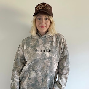 May include: A brown trucker hat with gold text reading "Sundays are for Football and Jesus". The hat is worn with a camouflage print hoodie with the words "faith over fear" in black.