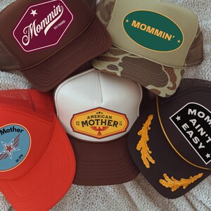 May include: A collection of trucker hats with various designs and text. One hat features "Mommin' So Hard" in a burgundy and white design. Another displays "MOMMIN'" in green. Other hats say "American Mother" and "Mom Ain't Easy".