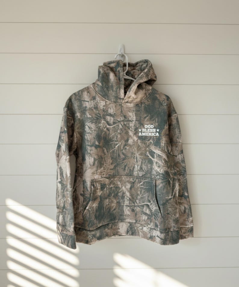 May include: Camouflage hoodie with a front pocket and a hood, hanging on a white wall. The text "GOD BLESS AMERICA" is printed on the upper right chest area of the hoodie.