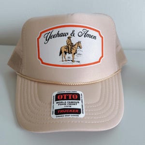 Puede incluir: Gorra trucker color beige con un gr&aacute;fico "Yeehaw & Amen" que muestra a un vaquero a caballo. La gorra tiene una parte trasera de malla y un detalle de cuerda en el borde. Tiene un cierre a presi&oacute;n duradero y una etiqueta OSFM.