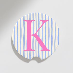 May include: Round car coaster with a cream background and blue vertical stripes. A large pink letter "K" is centered on the coaster. The coaster has a small notch for easy removal from a car cup holder.