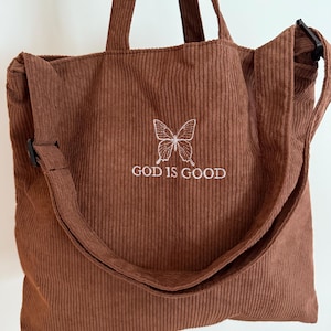 Puede incluir: Una bolsa de mano de pana marrón con una mariposa bordada en blanco y el texto "GOD IS GOOD".