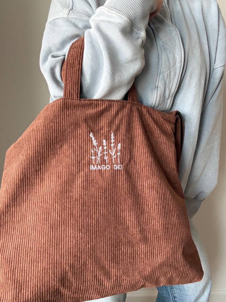 Puede incluir: Una bolsa de mano de pana marr&oacute;n con flores bordadas en blanco y el texto "IMAGO DEI".